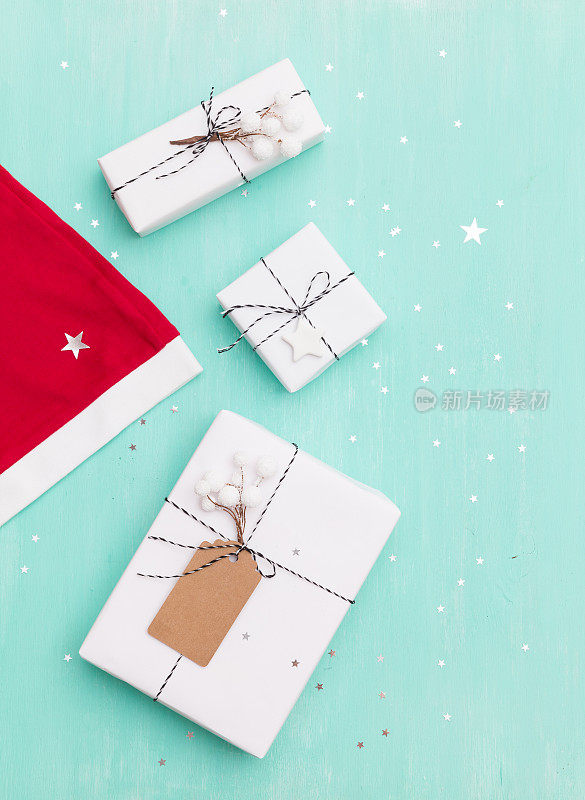 Top view on Santa's red hat and Christmas gifts wrapped in white paper and decorated with twine and tag on turquoise wooden background with闪亮的银色星星